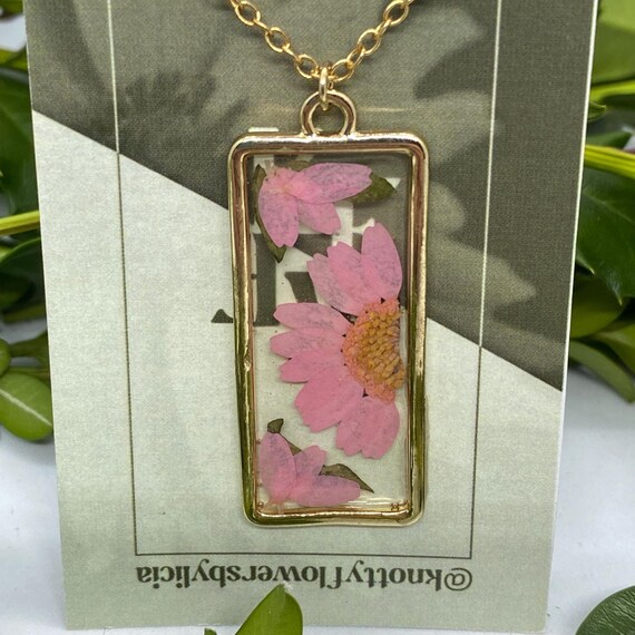 Handmade resin necklace with a bright pink daisy flower as the centerpiece, half of the daisy is in the middle of the right side while on the left petals with green leaves are in the corners framing the half daisy