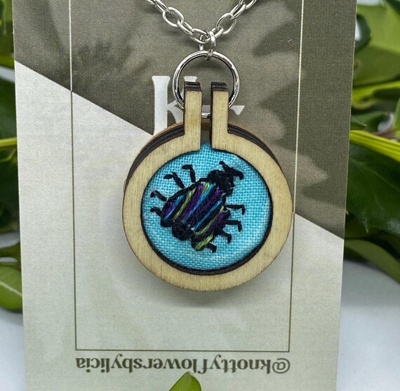 Hand stitched embroidered beetle bug with purple, blue, and green vertical stripes on its back and 4 legs on each side.It has large antennae on its head. The bug is on a sky blue background